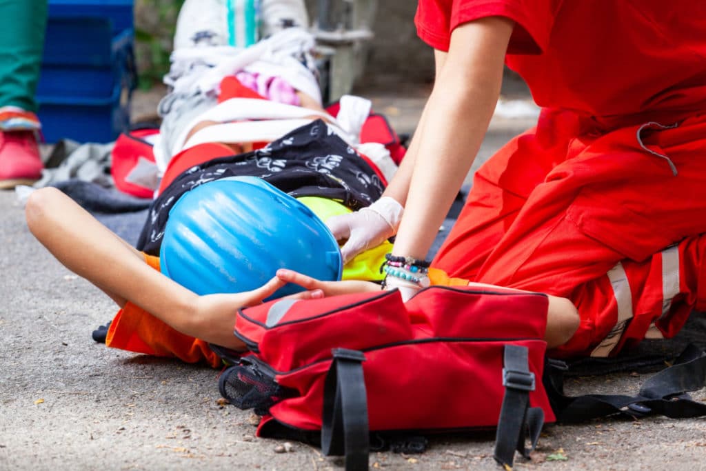 une personne allongée avec un casque de chantier bleu se voit prodiguer des soins par un secouriste en combinaison rouge.